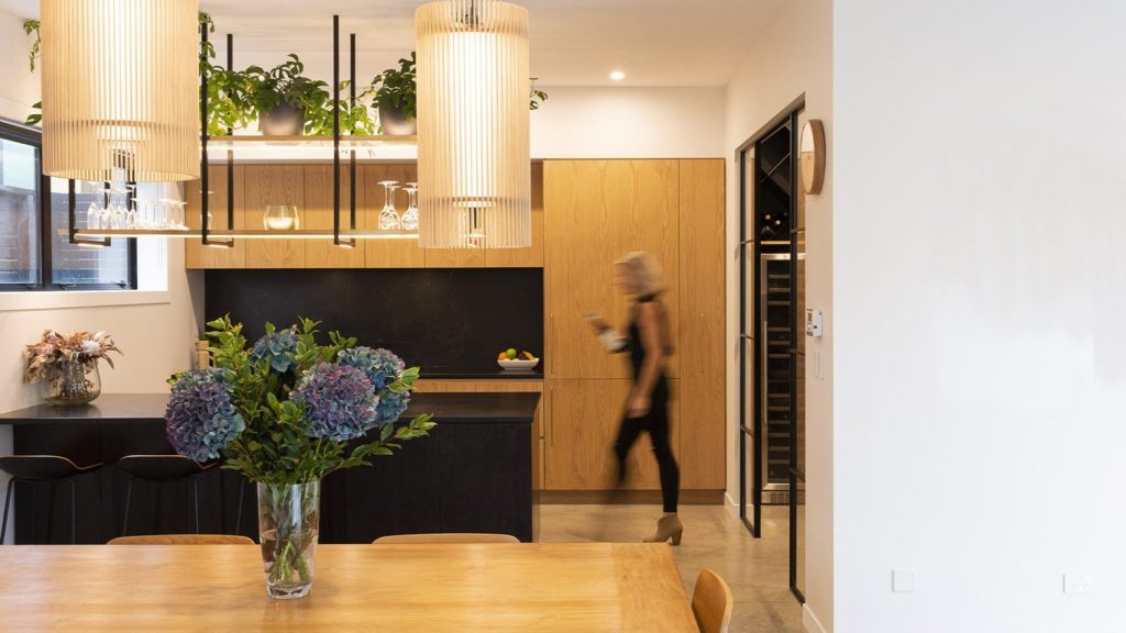 Modern Westmere kitchen with custom American oak cabinetry, black island, and hanging shelving with greenery, woman walking through blurred in foreground