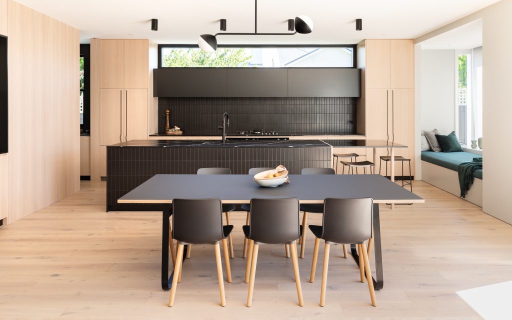Full view of bespoke kitchen renovation project - Mt Eden kitchen island by Fluid Interiors, featuring warm oak veneer cabinetry, honed black Eternal Marquina Suede stone benchtops, custom black powder-coated steel breakfast bar with oak round legs, and adjacent dining table.