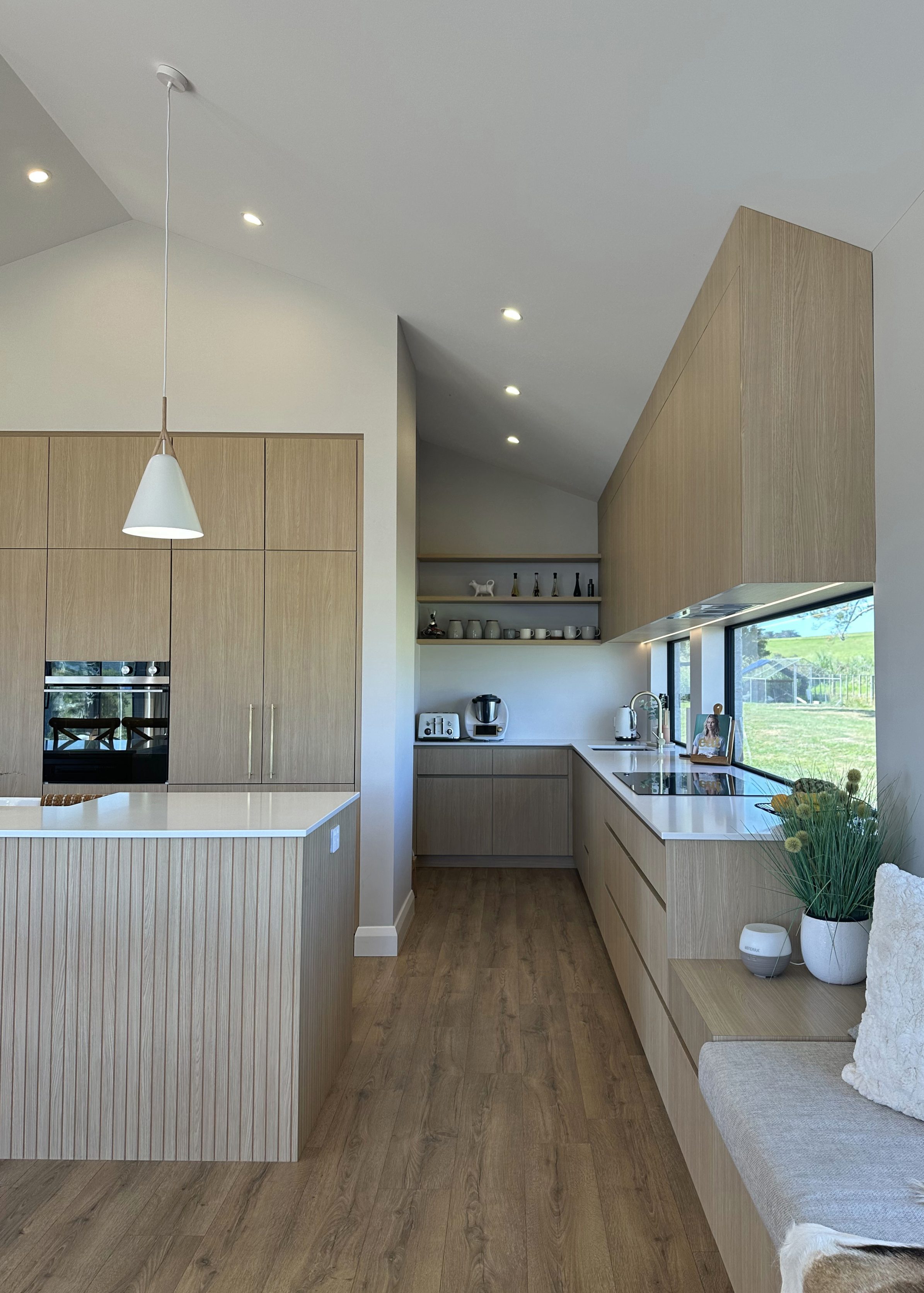 View from bench seat showing back of profiled Aged Ash Melteca kitchen island leading to hidden scullery in Kaukapakapa home by Fluid Interiors.