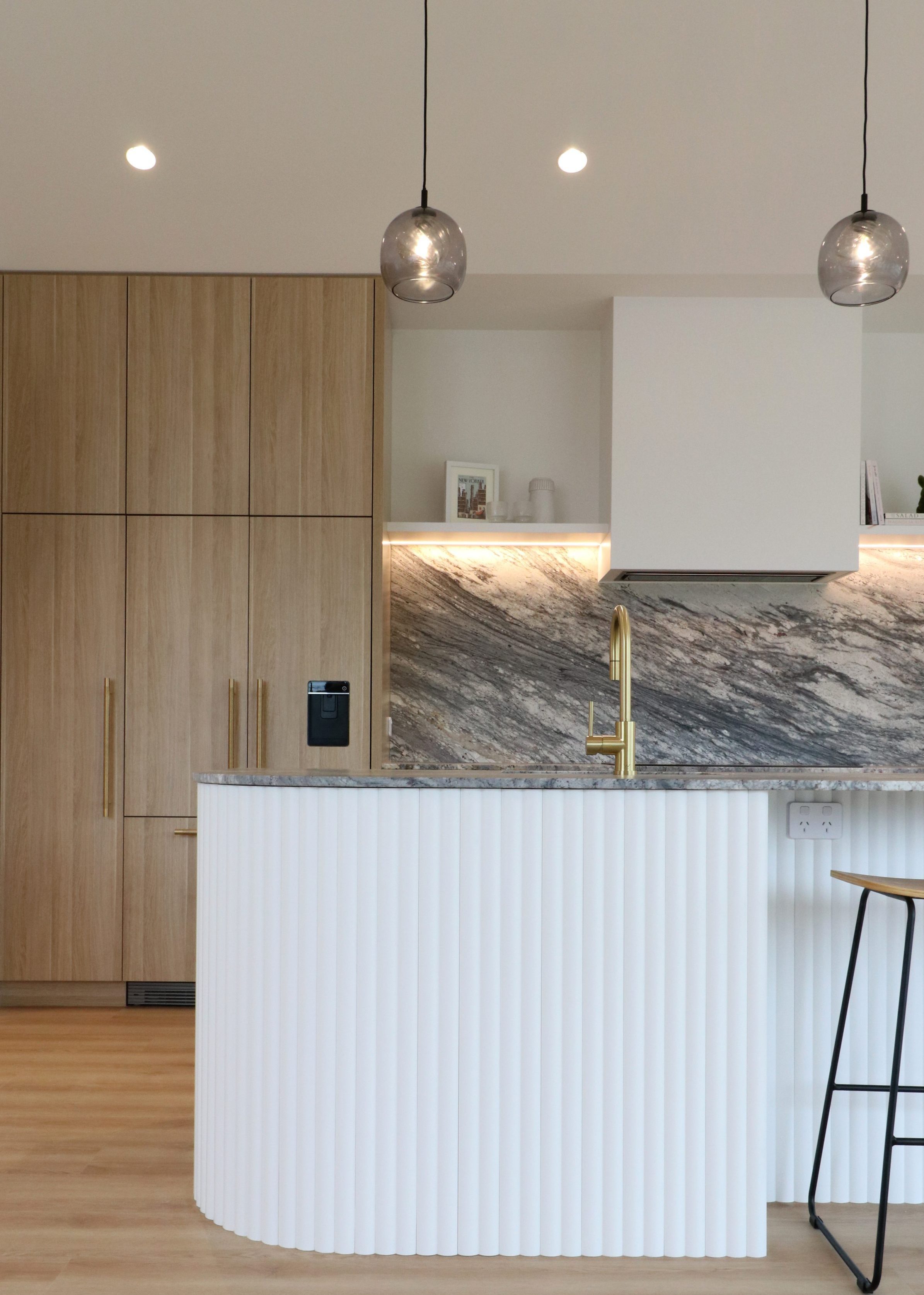 Portrait view of curved Genia-clad island, integrated fridge with oak panels, and open shelving beside custom rangehood