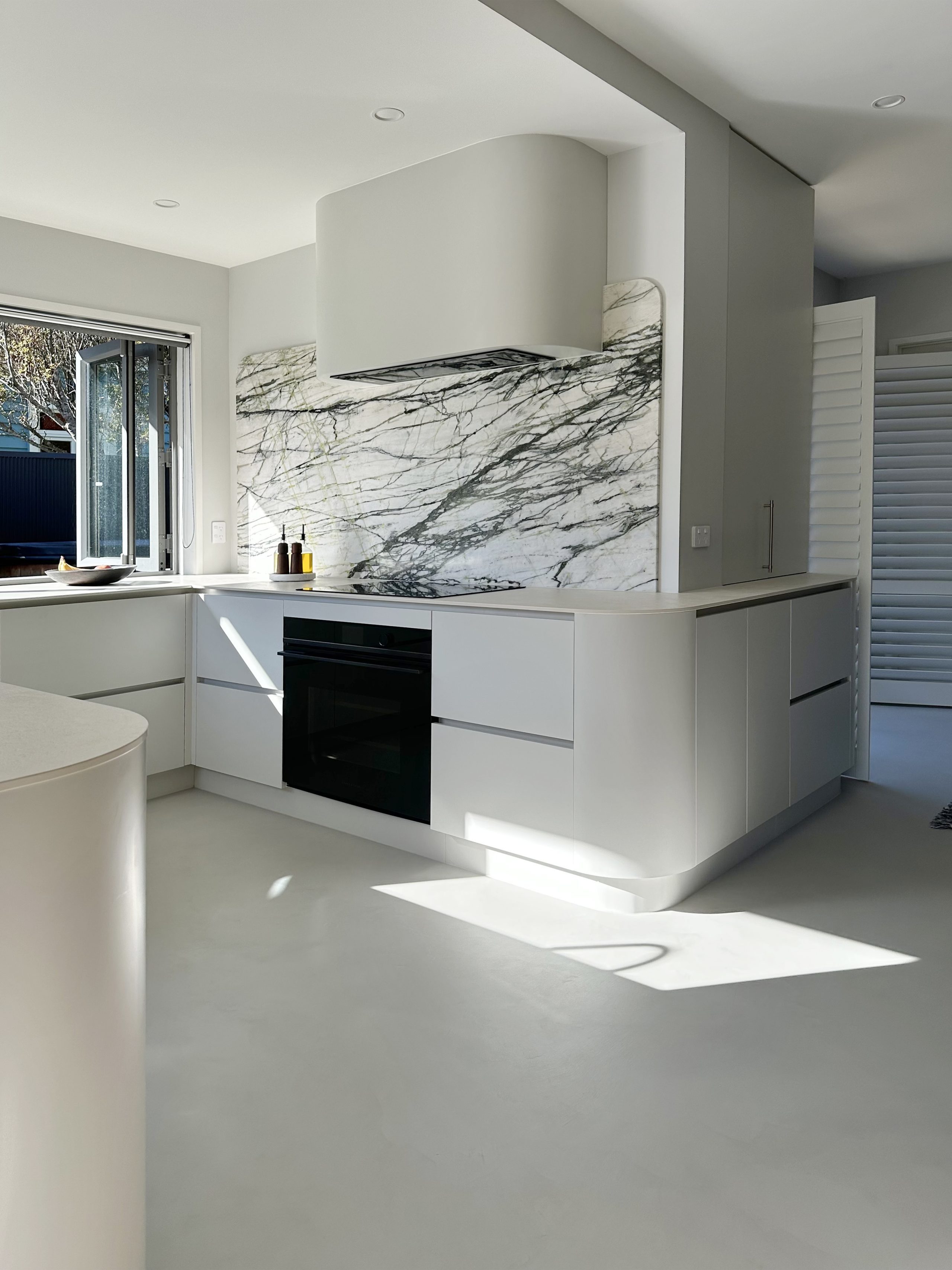 Melteca Baikal Natural base cabinetry with tall bi-fold unit, custom curved rangehood, and Calacatta Verde marble splashback in the Bayswater kitchen by Fluid Interiors.
