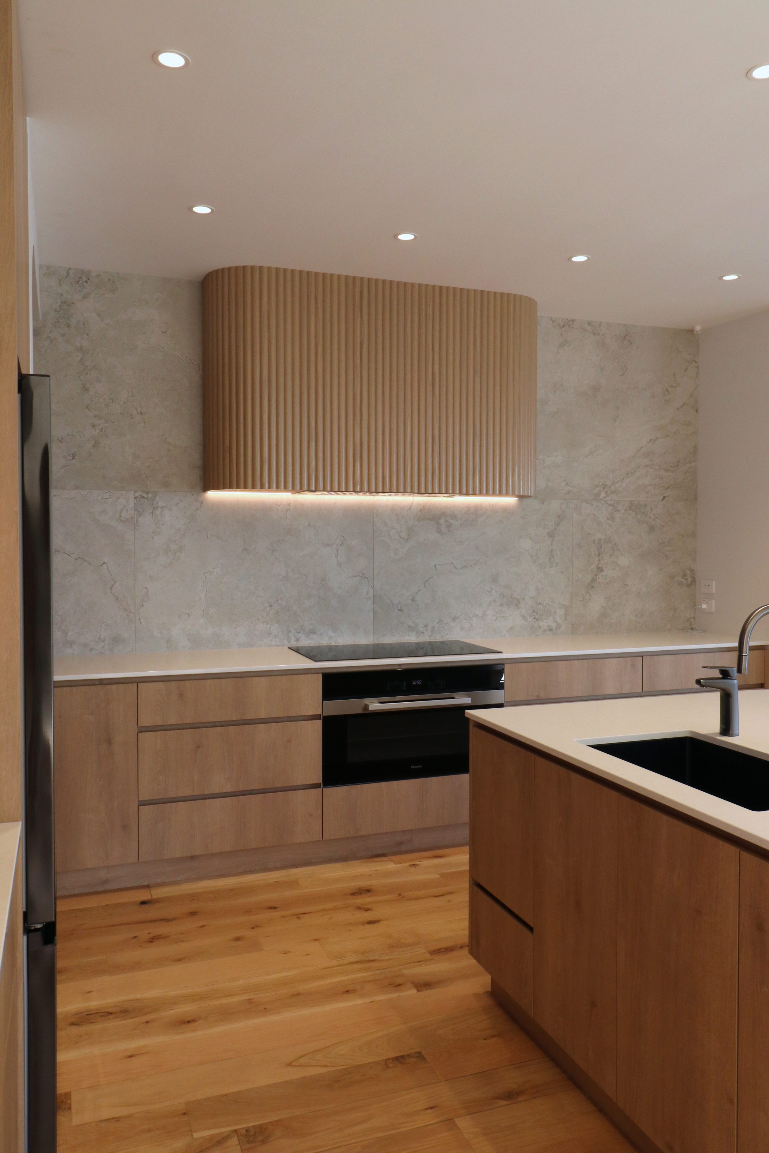 Angled view of custom fluted oak rangehood with integrated extractor, framed by textured tiles and soft LED lighting in a modern Auckland kitchen.