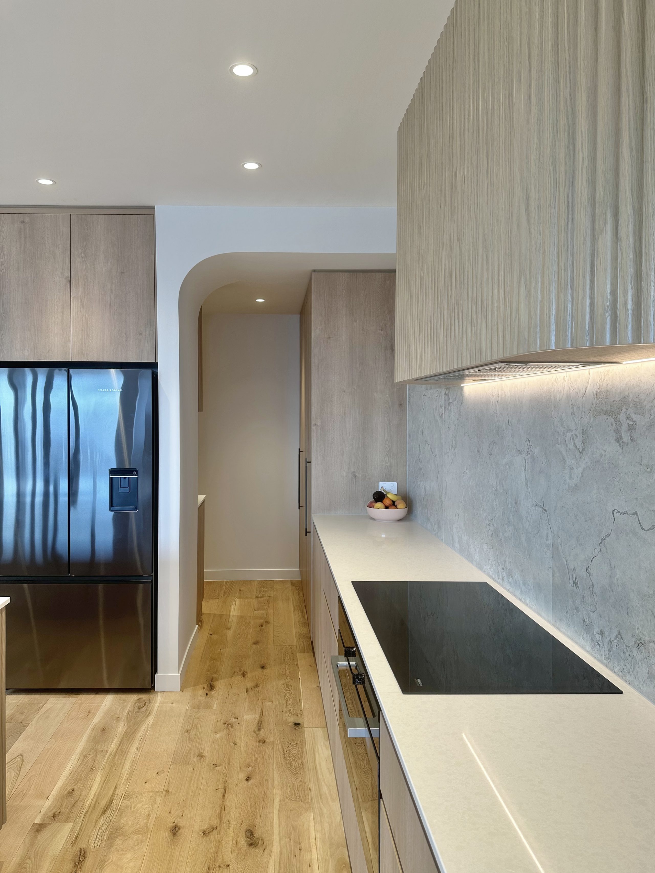 View looking past the fluted oak rangehood through a curved archway entrance to a functional scullery with ample storage in a bespoke Auckland kitchen.