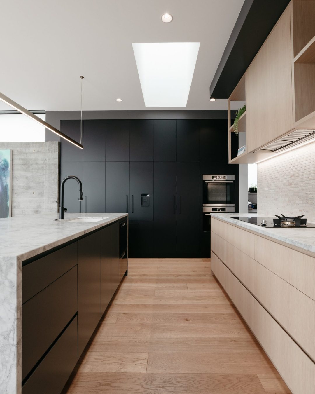 Straight-on portrait view of tall matte black cabinetry with integrated appliances, American oak veneer cabinetry, and honed Super White granite benchtops in Herne Bay kitchen by Fluid Interiors.