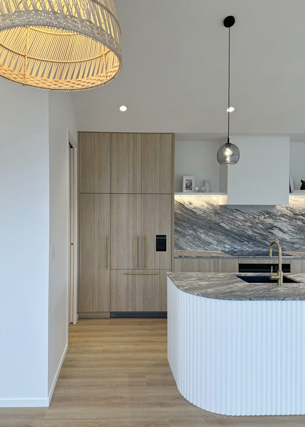 Portrait view of curved Genia island with Sensa granite top, integrated fridge with oak panels, custom rangehood and open shelving