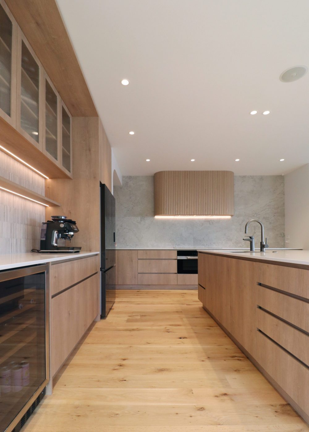 Wide view of fluted oak rangehood with integrated extractor, large-format tile splashback, and adjacent coffee nook with terrazzo tiles and fluted glass.