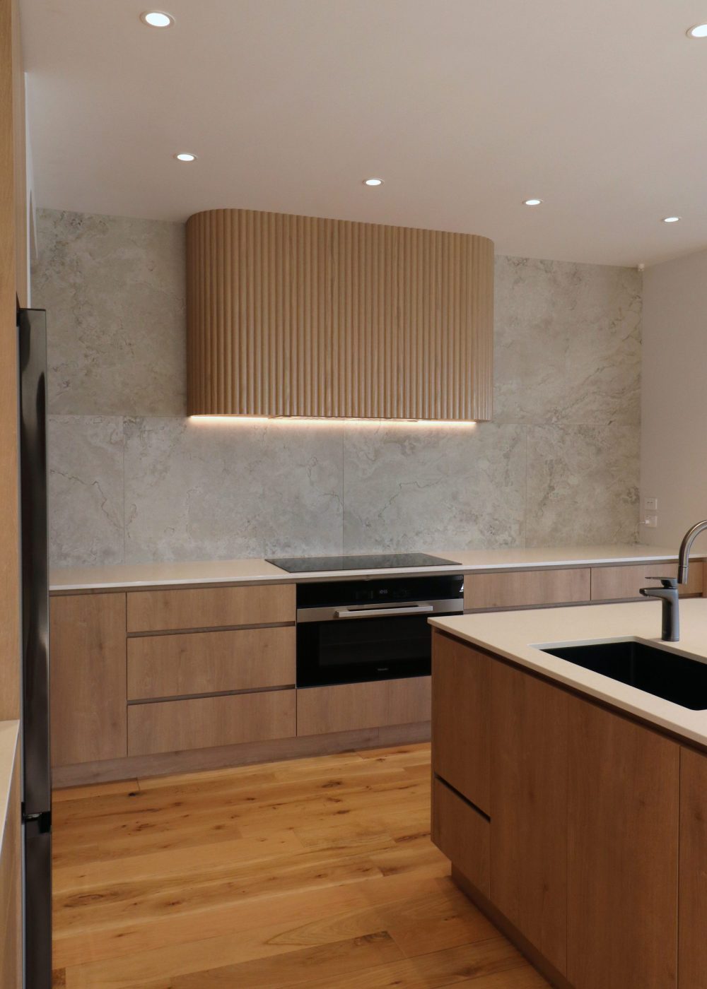 Angled view of custom fluted oak rangehood with integrated extractor, framed by textured tiles and soft LED lighting in a modern Auckland kitchen.