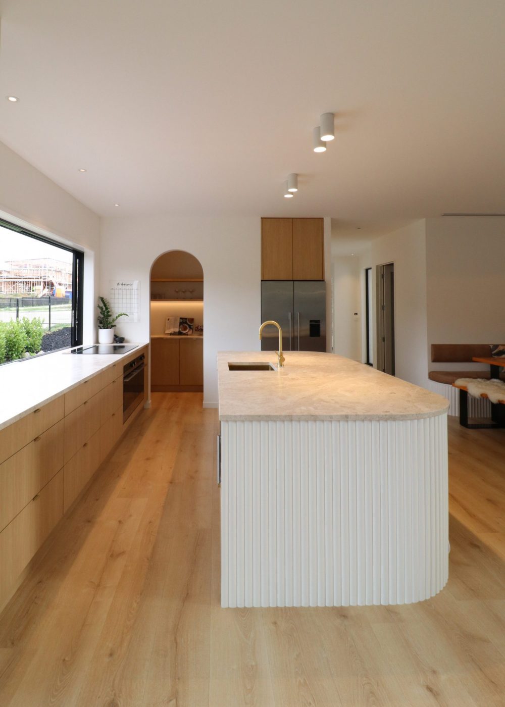 Portrait view of Wainui kitchen with curved Genia panelling island, oak-toned Melteca Puregrain cabinetry, and arched scullery entry framed by soft natural textures and Marble SaSaab benchtops.