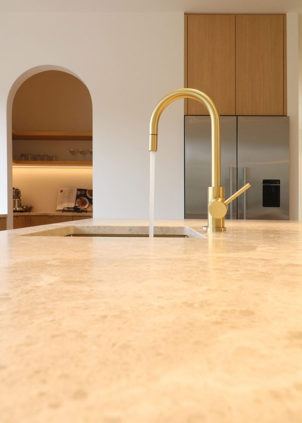 Close-up of running tap in bespoke Wainui kitchen by Fluid Interiors, with curved scullery entrance and integrated fridge visible in background. Features aged ash Melteca Puregrain cabinetry and Marble SaSaab benchtops.