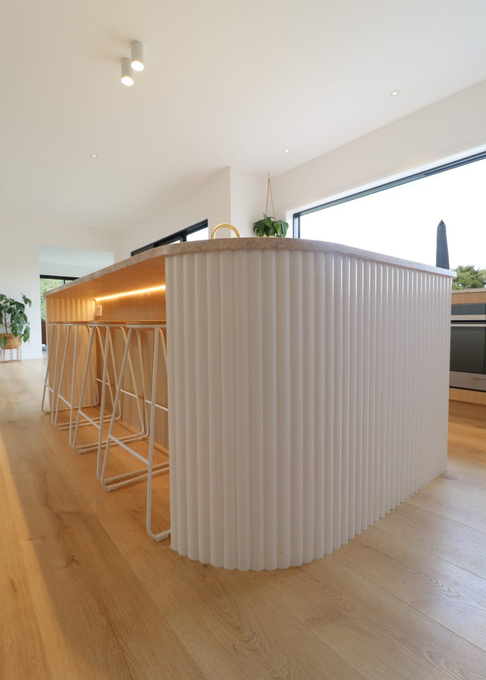 Close-up of curved kitchen island in Wainui home by Fluid Interiors, showing under-bar LED lighting, fluted Genia panelling, and honed Marble SaSaab benchtop.